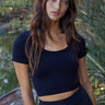 Model wearing the Tamara Top in black, featuring a soft ribbed knit fabric, scoop neckline, short sleeves, and fitted cropped silhouette, styled with a matching black skirt.