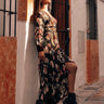 A person wearing a floral maxi dress with long sleeves, styled with a belt, and black boots, standing in a narrow alley with buildings in the background.