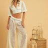 A female model wearing a white eyelet crop top and matching pants, standing next to a basket and a pot, on a beige background.