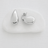 Polished silver teardrop Trena stud earrings resting on a white stone against a neutral fabric background.