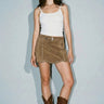 Woman wearing the Yagu Skirt, a suede-look western-inspired mini skirt featuring silver stud detailing, adjustable side buckle, high-rise waist, and rich camel color styled with a white tank top and cowboy boots.
